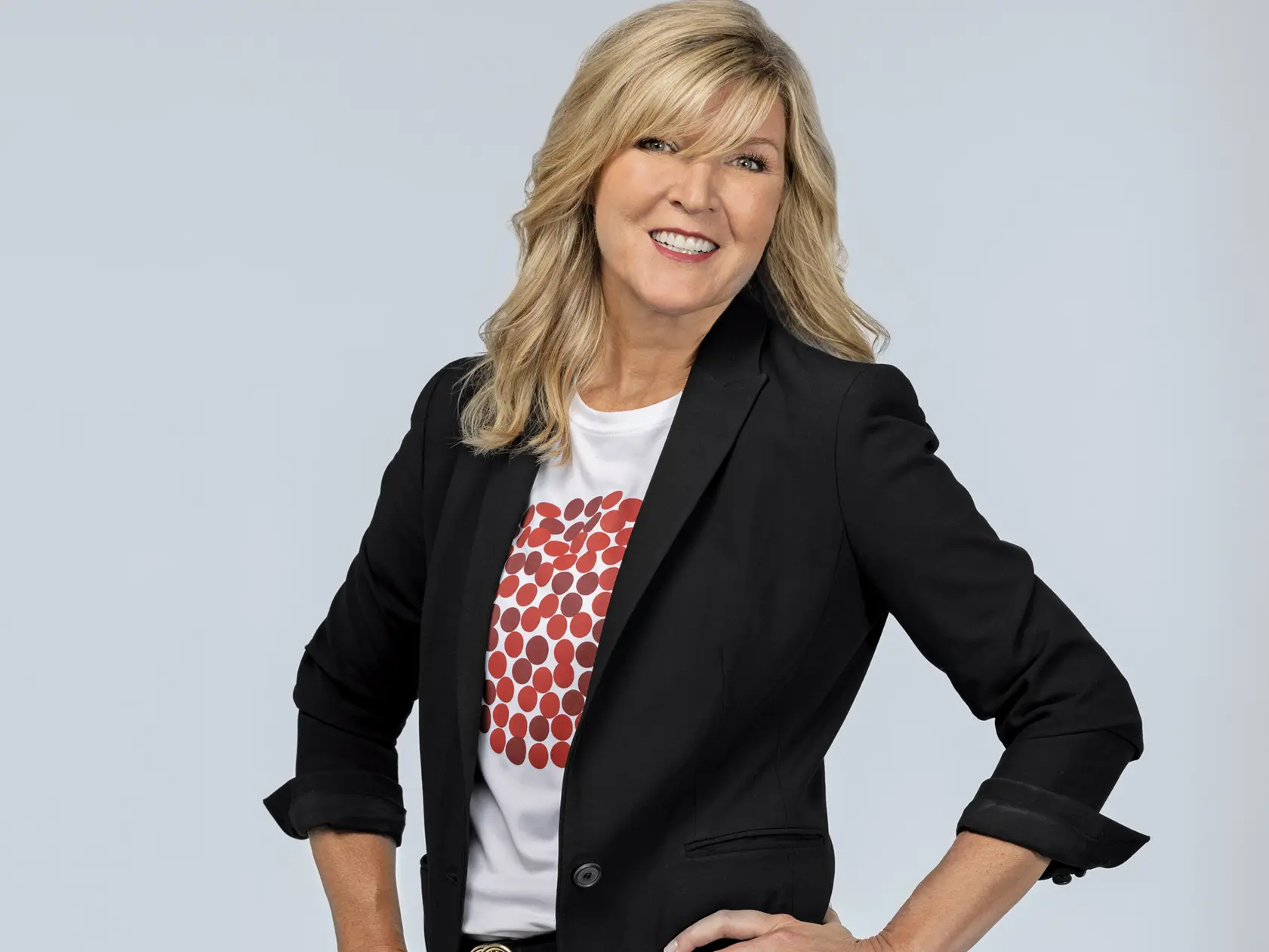 A white woman wears a pepperoni pizza T-shirt, under a black suit jacket, with black pants. She has blond hair, blue eyes, a big smile and stands confidently with her hands on hips. 