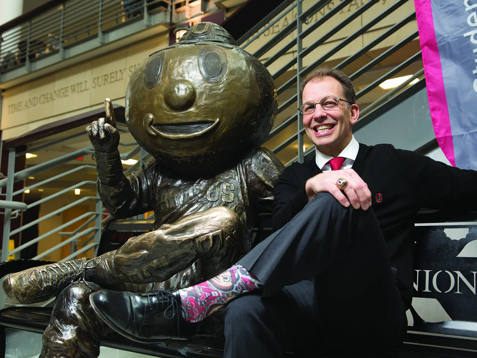 A man smiles as he sits on a bench next to a bronze statue of Brutus. Their leg-crossed poses and smiles are similar.