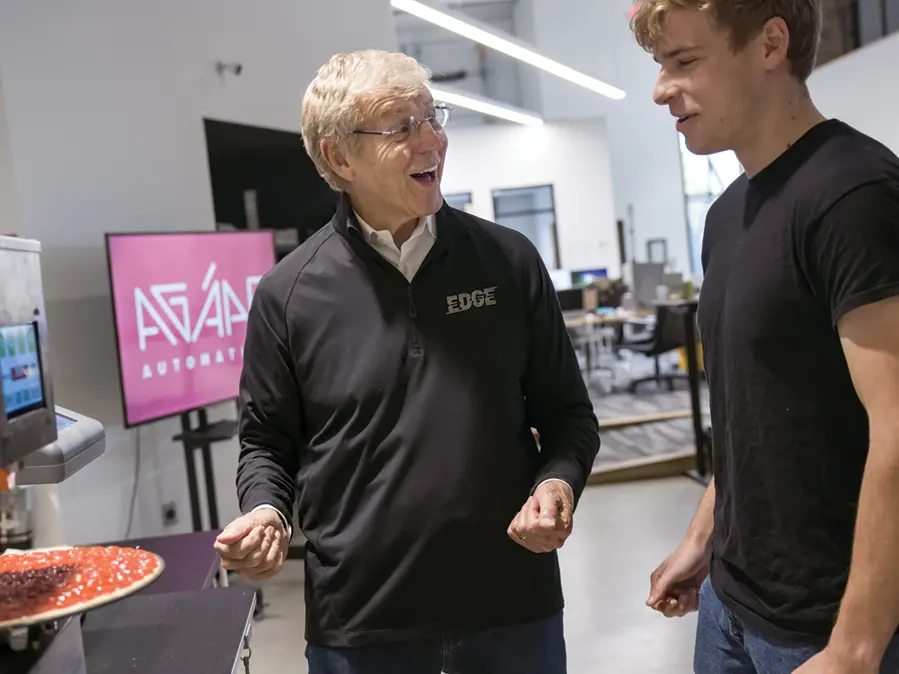 A college student watches an automatic pizza saucing machine as an older man talks to him excitedly.