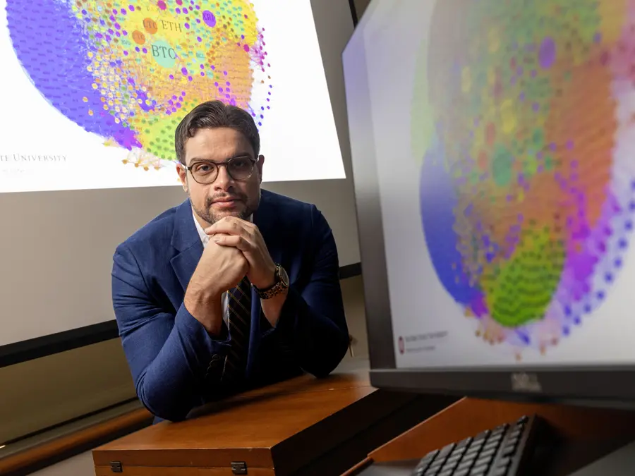 In a classroom setting, a man wearing a suit and glasses looks confident and thoughtful as he leans on a desk between colorful charts.