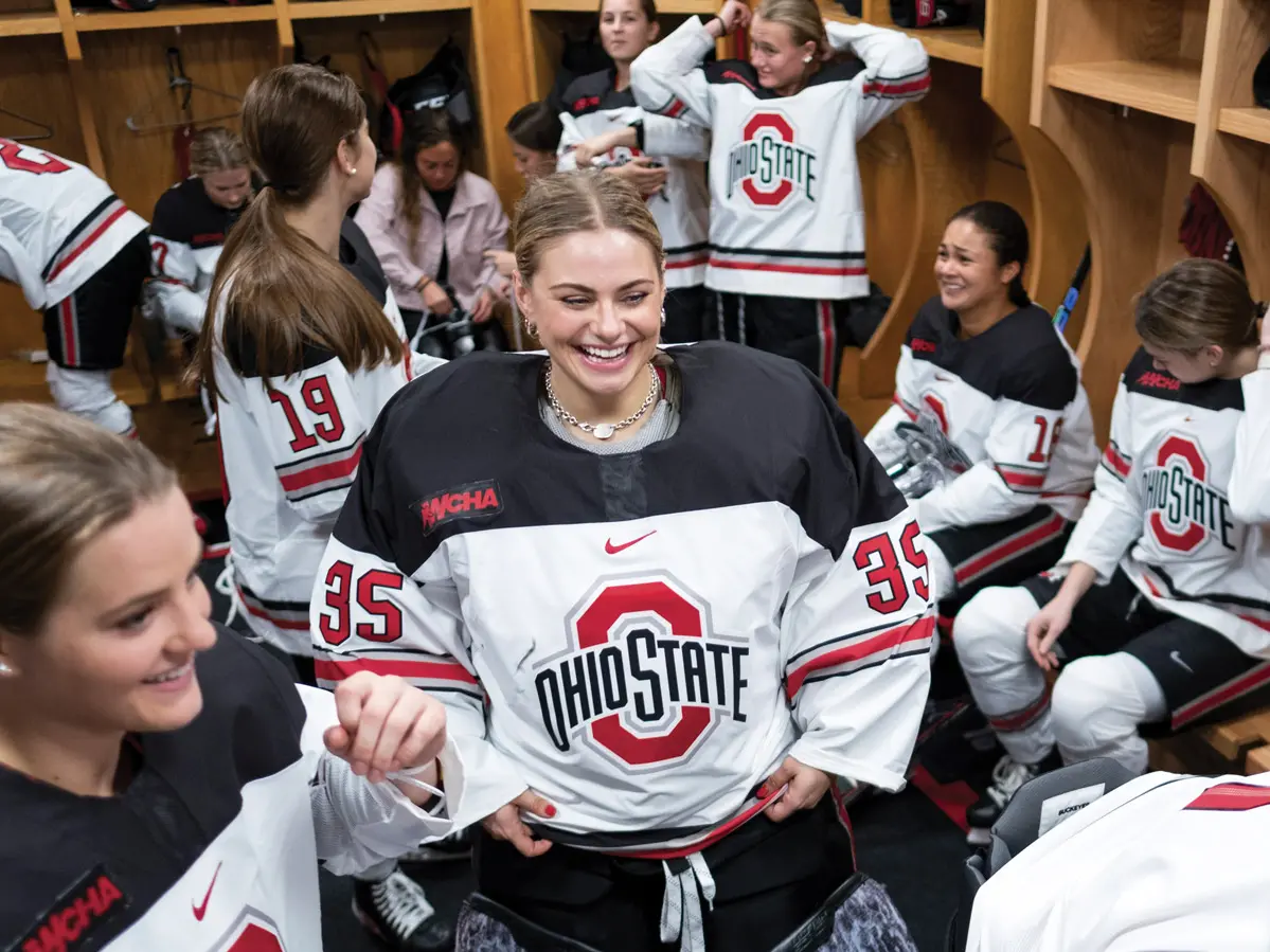 In a crowded locker room, young women in ponytails and Ohio State hockey uniforms laugh and talk in groups.