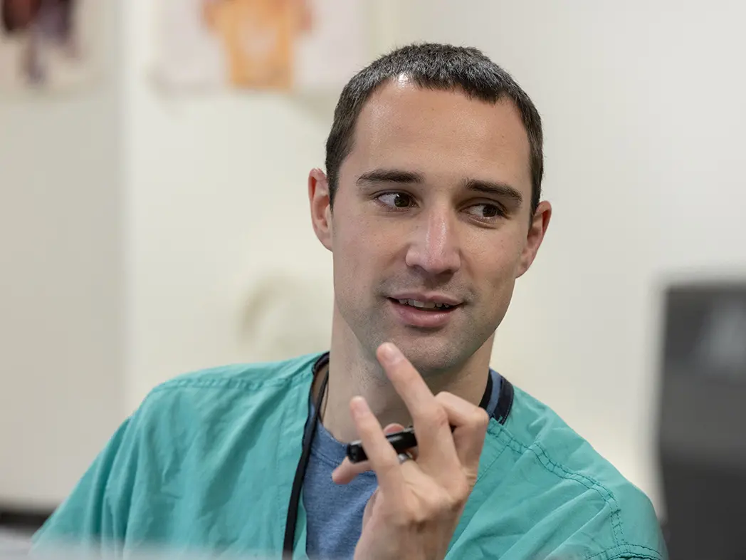 A white man with short dark hair and dark eyes smiles and twirls a pen as he looks at someone off camera. He’s wearing green scrubs and a lanyard.