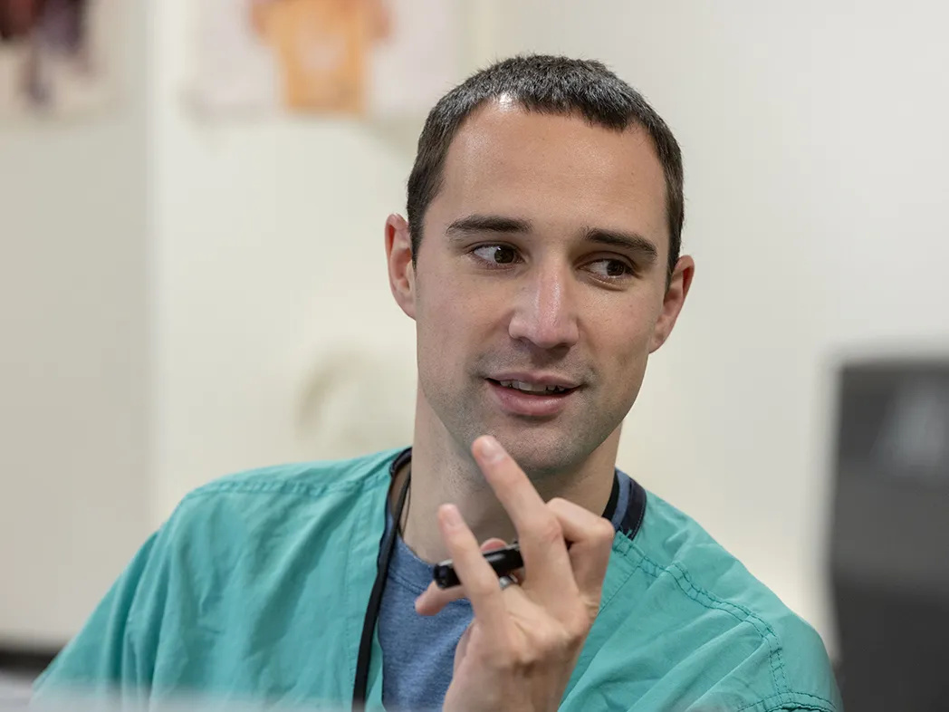 A white man with short dark hair and dark eyes smiles and twirls a pen as he looks at someone off camera. He’s wearing green scrubs and a lanyard.