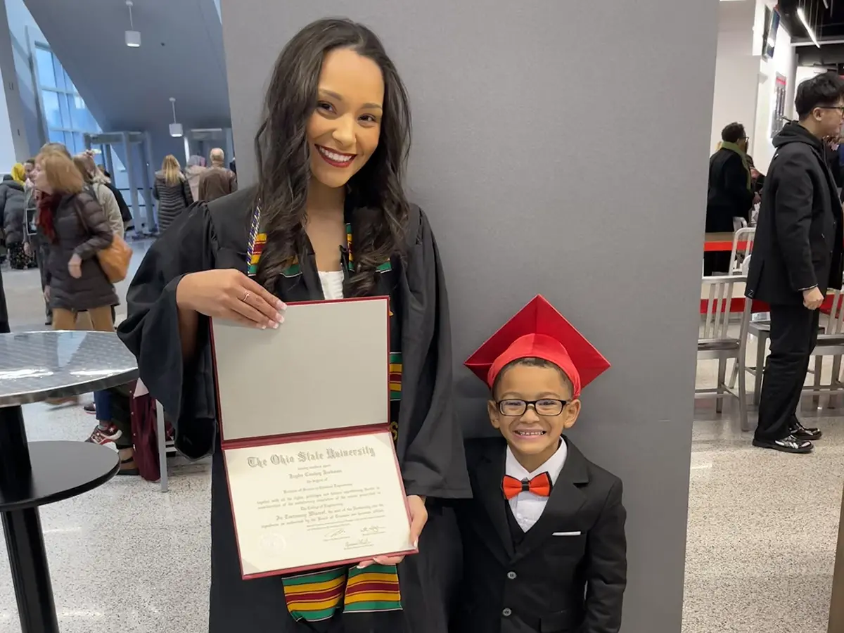 A young black woman in a graduation robe smiles brightly as she shows her diploma, and her son, who seems to be no older than 5 or 6, grins. He’s wearing a little suit with a scarlet bow tie and graduation cap.