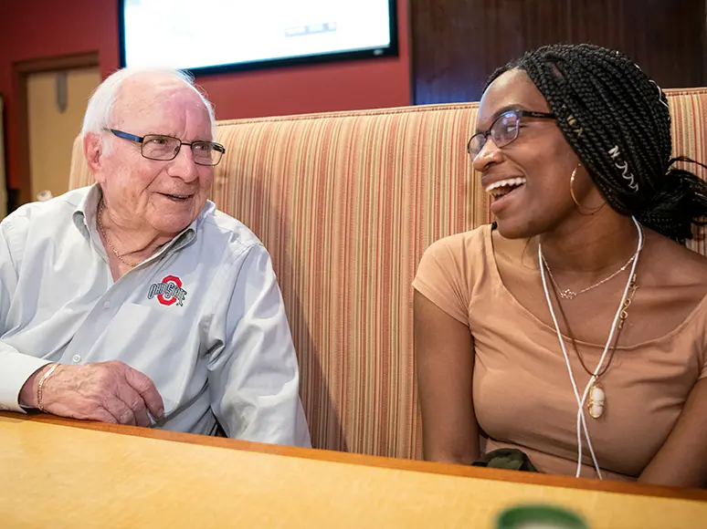 AN older white man and young Black female student sit in a booth. Both are smiling as they talk to each other.