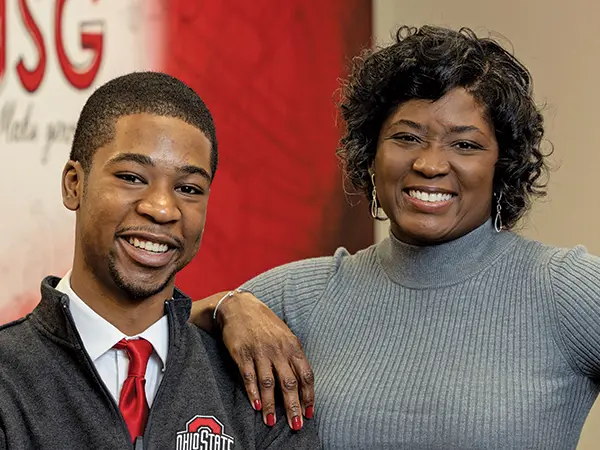 A young black man poses with his mom. He’s wearing a button down, red tie and Ohio State pullover, and she looks stylish in a gray dress. They both wear genuine smiles, as evidenced by the way their eyes crinkle.