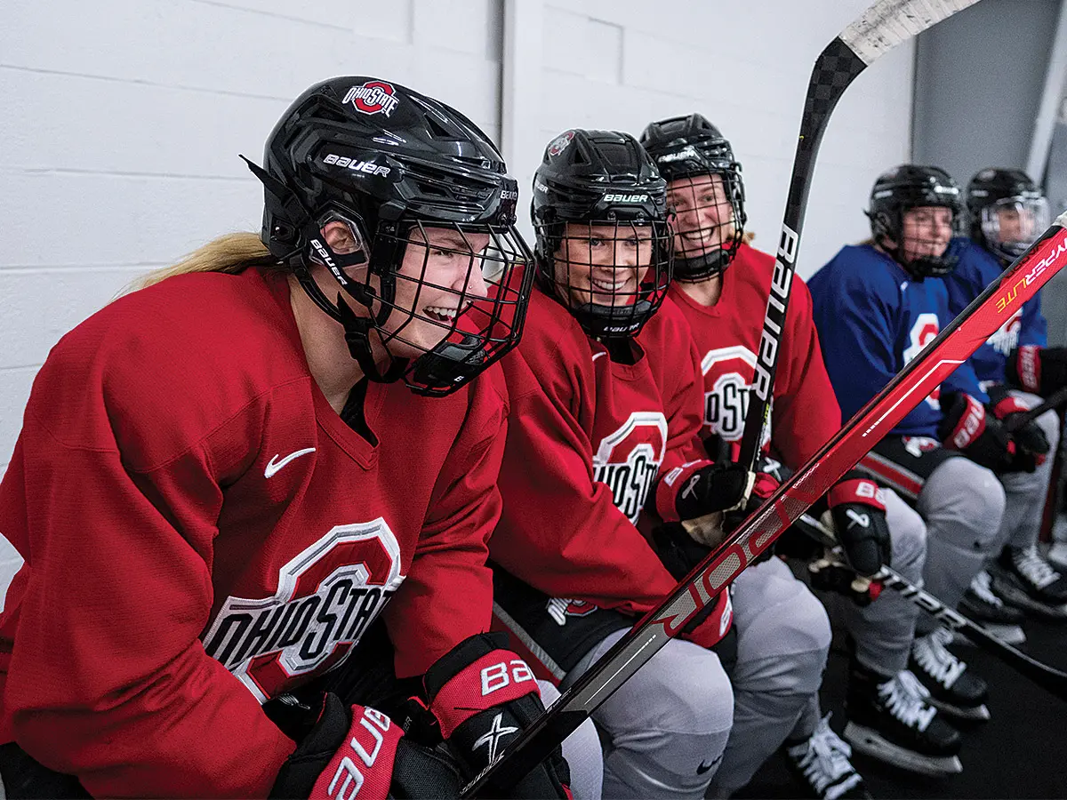 Sitting on a bench during practice, helmeted ice hockey players hold their sticks and laugh and talk while watching the ice (not shown in the photo). The first three wear red jerseys and the last two wear blue, but all have big Ohio State logos on the chest.