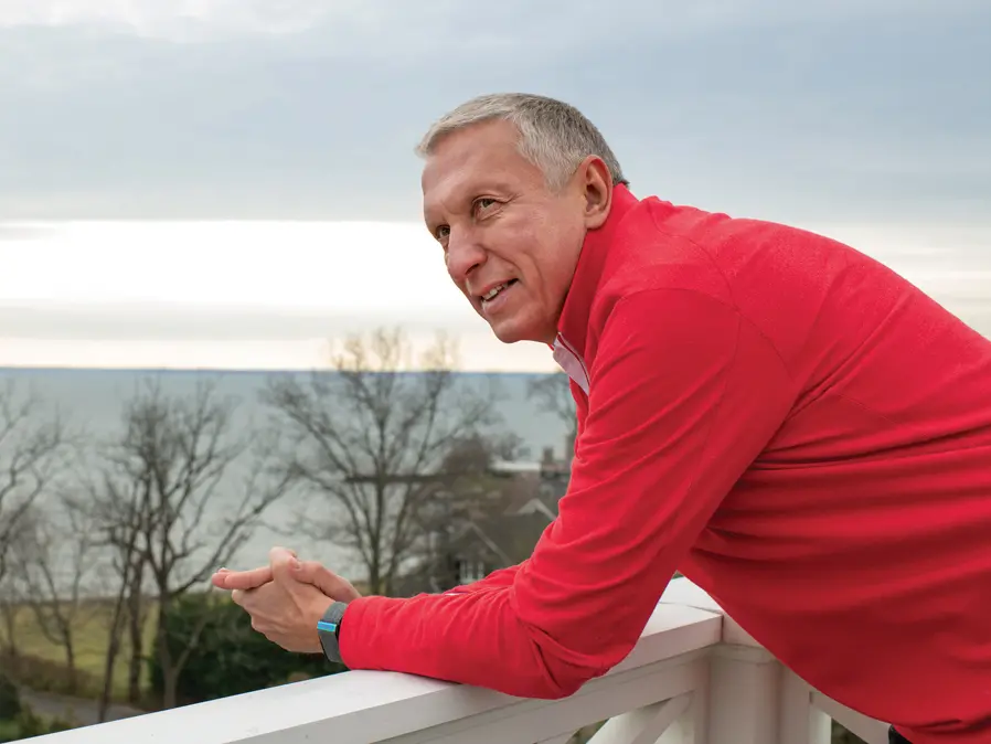 A white-haired man wearing a scarlet shirt leans on a railing on a roof-top porch with a view of the ocean. He has an interested, happy expression, as if he is looking, not at the pretty scene, but at someone talking next to him.