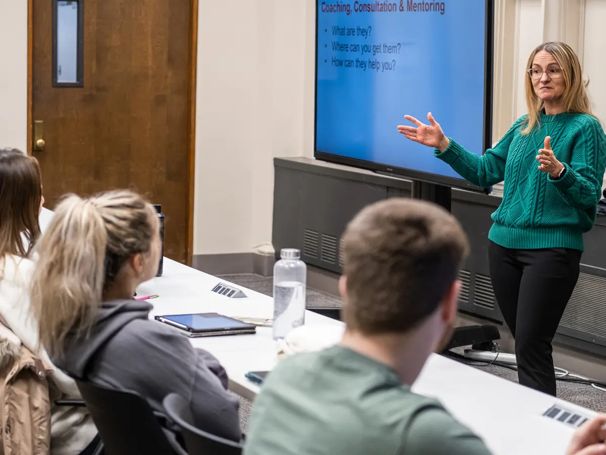 In an Ohio State College of Optometry class, four students sit at a long table at the front of a classroom watching a teacher define mentoring. The teacher, a blond white woman wearing glasses, has her hands spread wider than her body, as if encompassing all of the students in the room. A screen behind her says “Coaching, Consultation &amp; Mentoring—what are they?”
