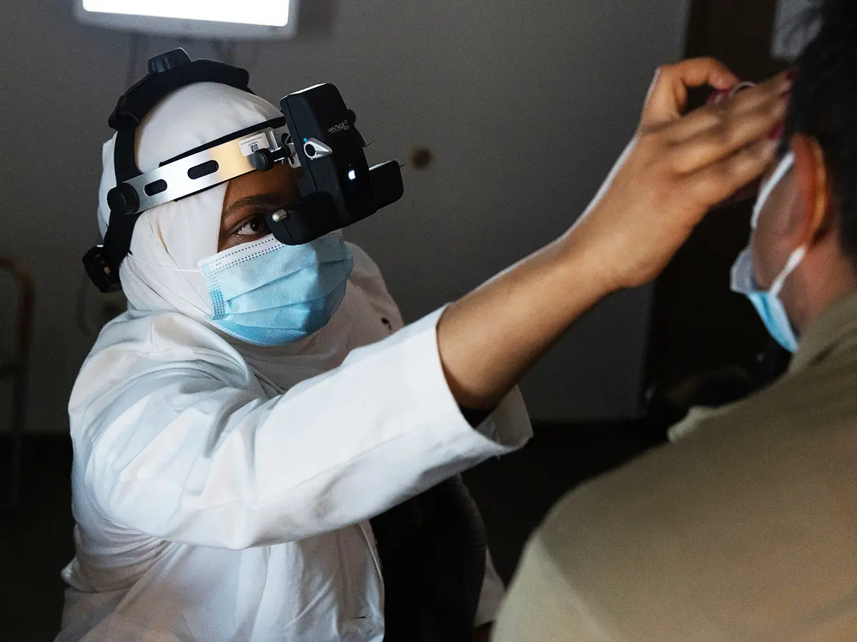 In a darkened room, an eye doctor examines a patient’s left retina by holding a small instrument in front of his eye and wearing a headset that is shiny metal and black plastic. The handheld part has a mirror that reflects light into the patient’s eye; the part on the doctor’s head, called a Binocular Indirect Ophthalmoscope, shows her a closeup view and provides the light.