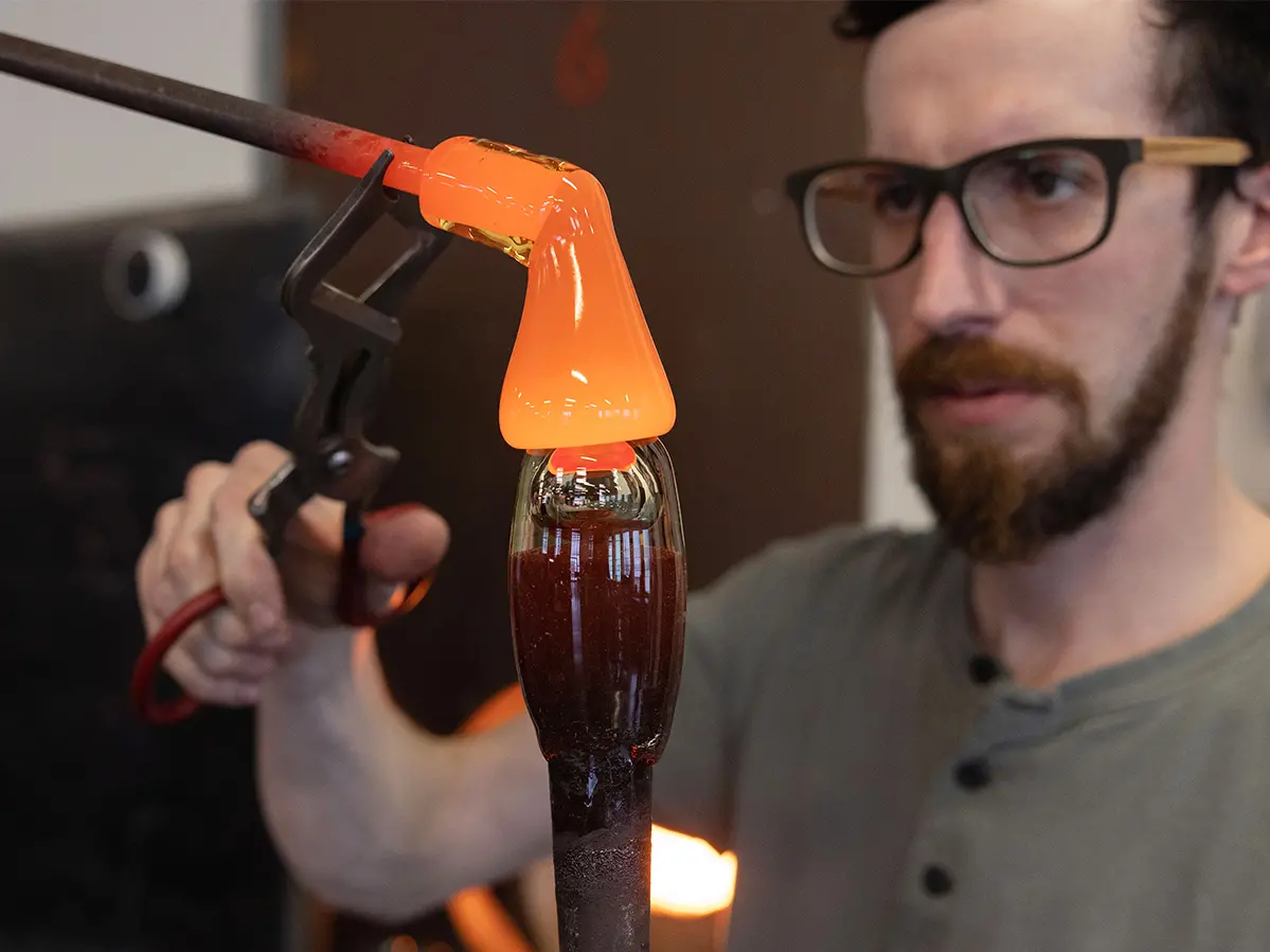 Artist John Sharvin uses metal tongs to steer molten glass connected to a blowpipe onto a cooled glass piece that is oblong. Sharvin, a white bearded man wearing glasses, seems incredibly focused as he takes care to create the effect he wants.