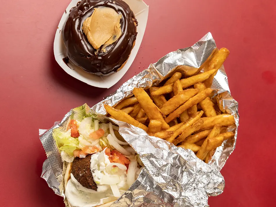 A tinfoil-wrapped gyro, with a falafel ball, lettuce, chopped tomatoes and tzatziki sauce, sits on top of a mound of French fries, each a little slimmer than a person’s pinky finger. Next the food is a doughnut topped with a thick coat of chocolate with a mound of peanut butter in the middle.