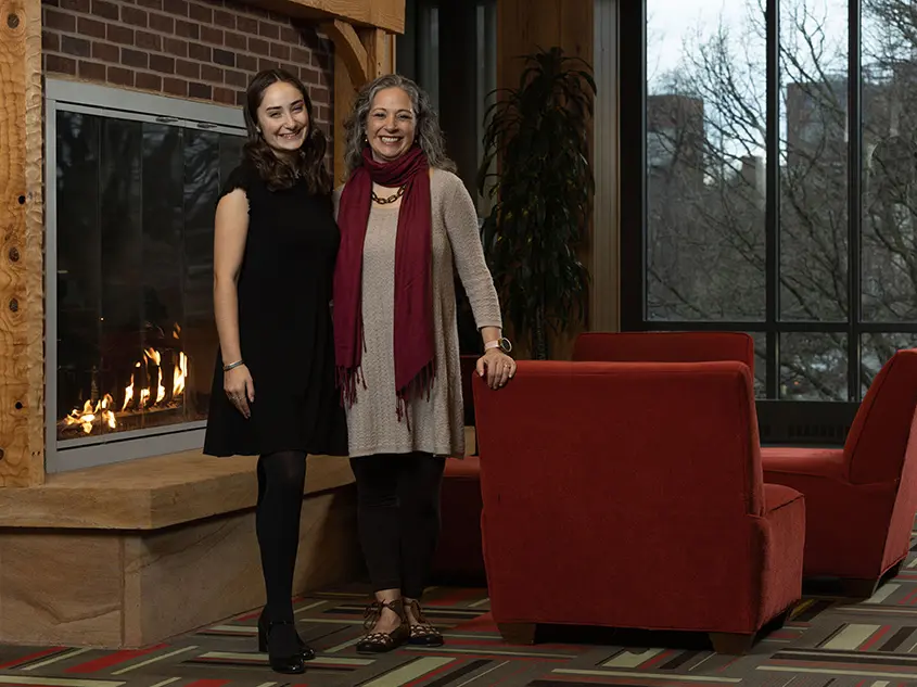 A young white woman poses with her mom in front of the fireplace at Longaberger Alumni House. They both wear dresses and big smiles, and have similarly wavy hair and stances.