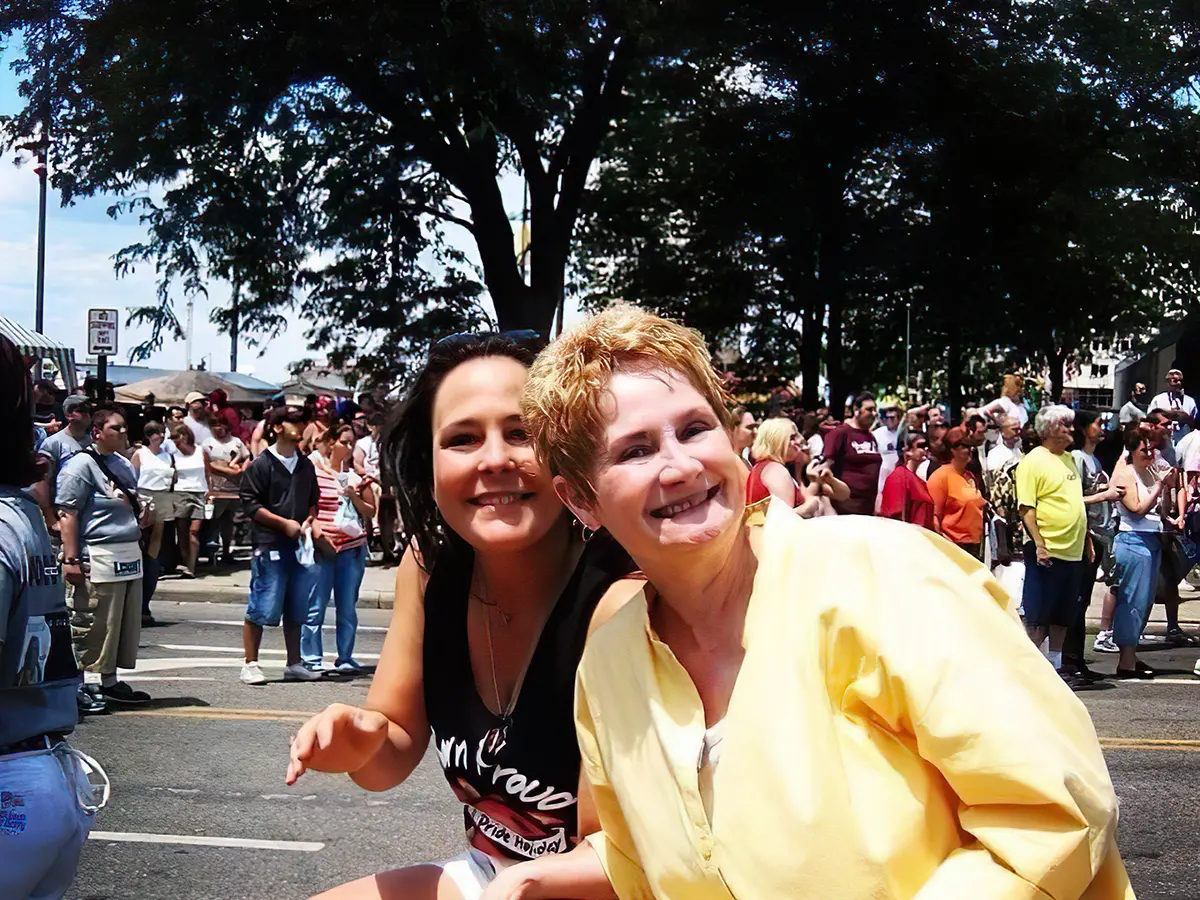 Two white women, one with short blond hair and the other with long dark hair, lean into the photo as parade-watchers line the street behind them. The women’s smiles say they are happy and proud.