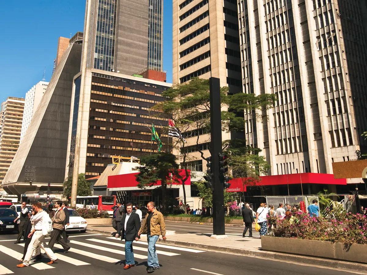 Skyscrapers line a street that has flower planters dividing the two directions of traffic. Pedestrians are shown crossing at a crosswalk. Bright blue sky peaks from behind the buildings.