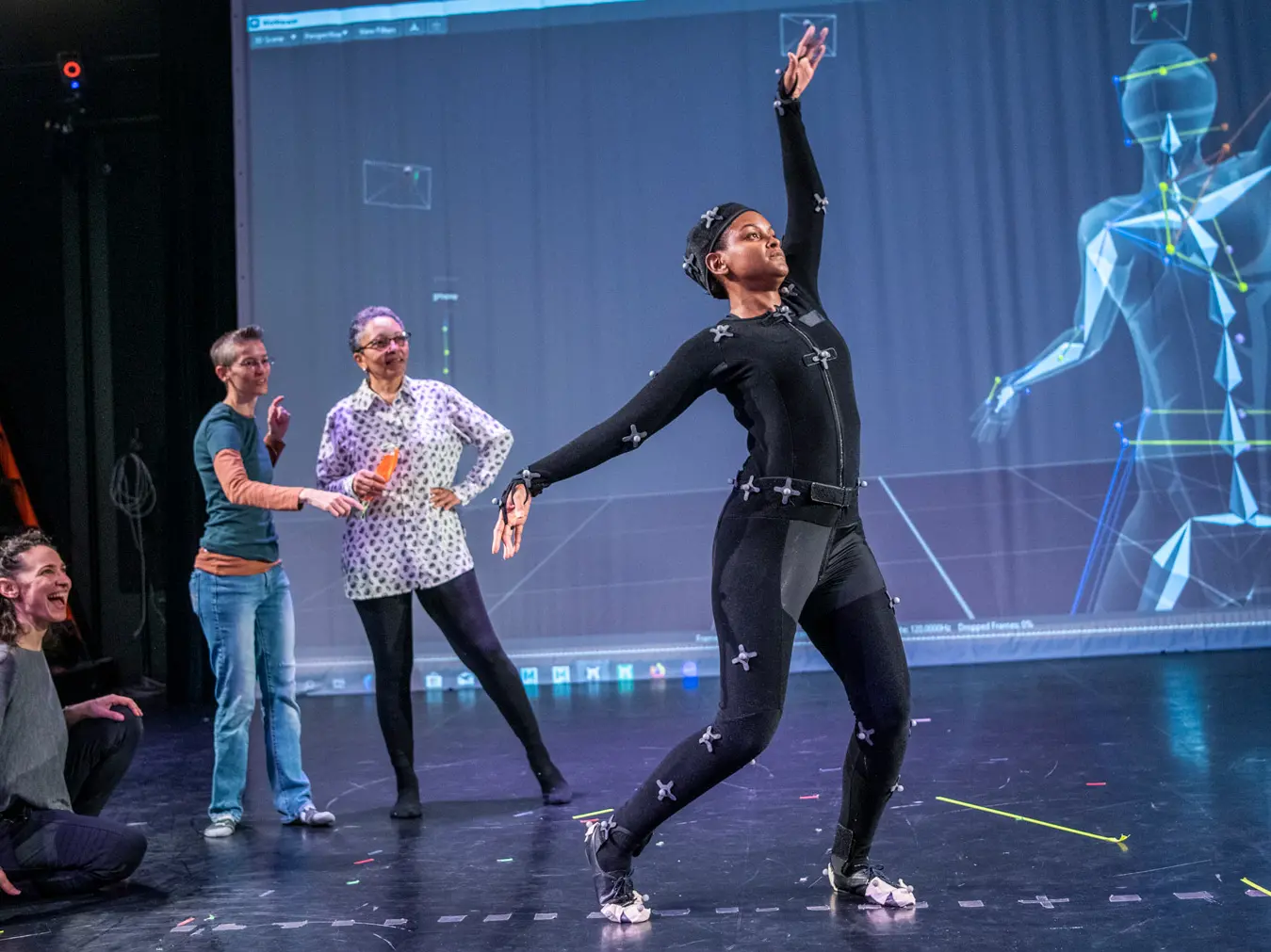 On a stage, a dancer in a black body suit and sensors steps forward and elegantly spreads her arms as three women behind her watch excitedly. A computer-generated geometrical illustration of the dancer highlights where her bones are as she moves.