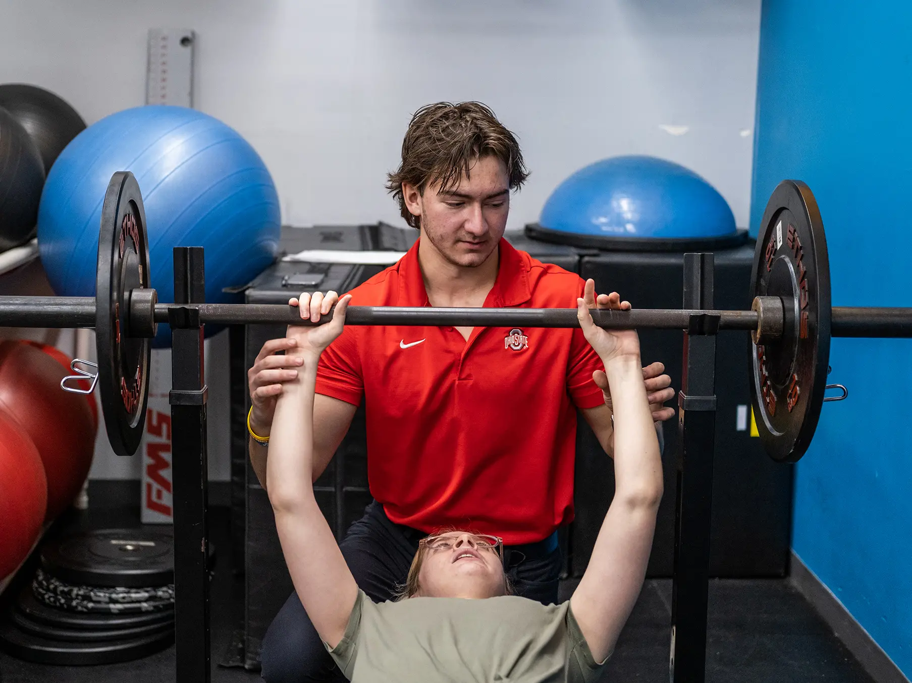 At a fitness gym, a young man with messy hair focuses on guiding a young woman getting ready to lift a weighted bar for a bench press. The man sits behind her, as she lies back on the bench and looks at the bar. He barely touches her wrists.  