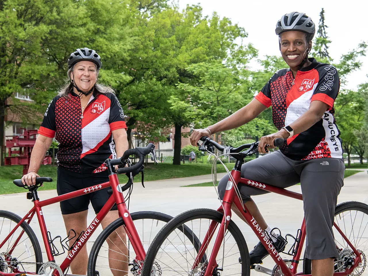 Two smiling women stand with their matching bicycles wearing matching Team Buckeye jerseys and bike helmets. On the left is Molly Ranz Calhoun, a tall white woman, and on the right is Provost Melissa Gilliam, a tall Black woman.
