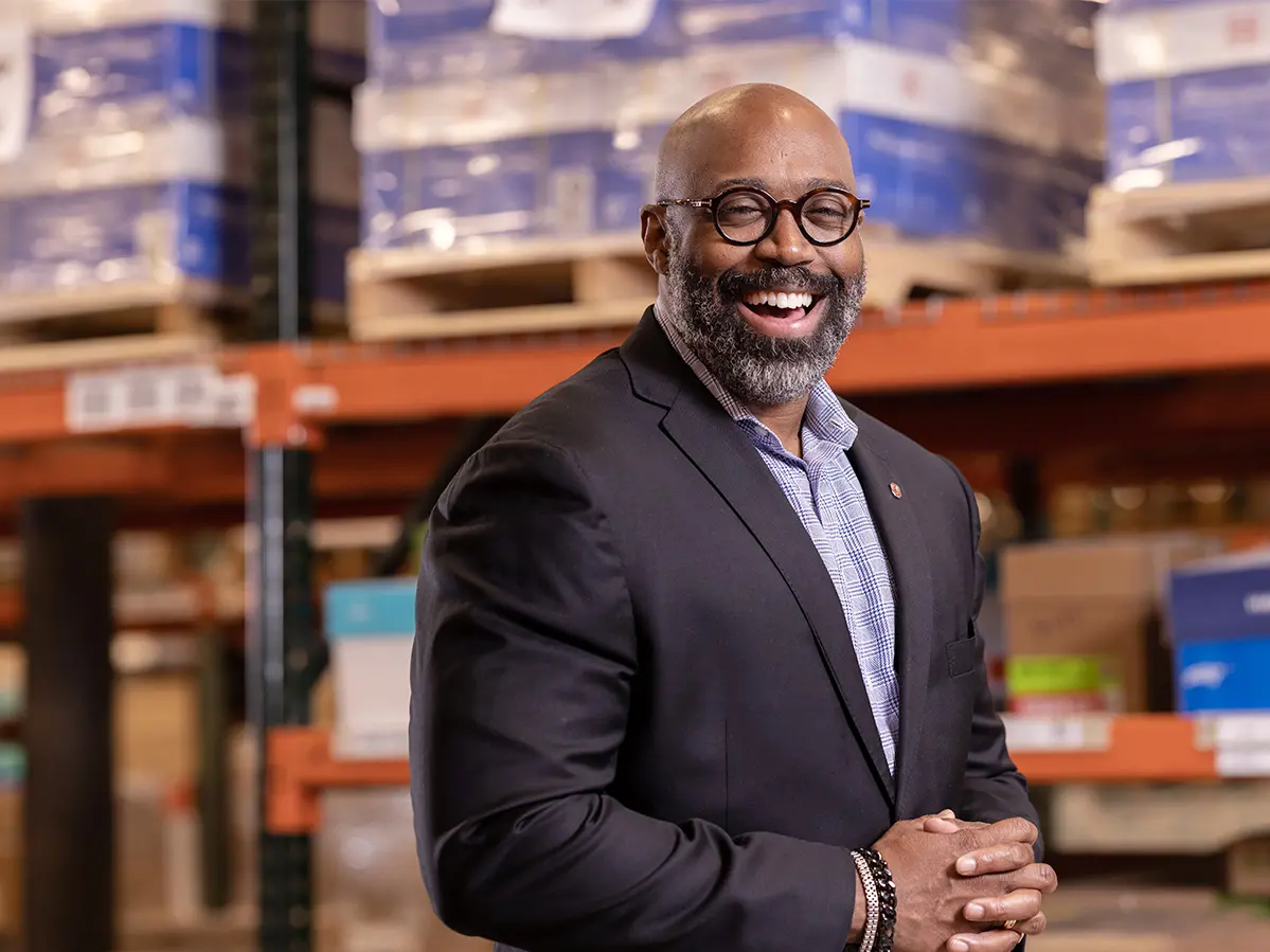 A bearded Black man whose head is shaved wears round glasses and a suitcoat and smiles in a wise way as he stands in a warehouse. His surroundings are out of focus.