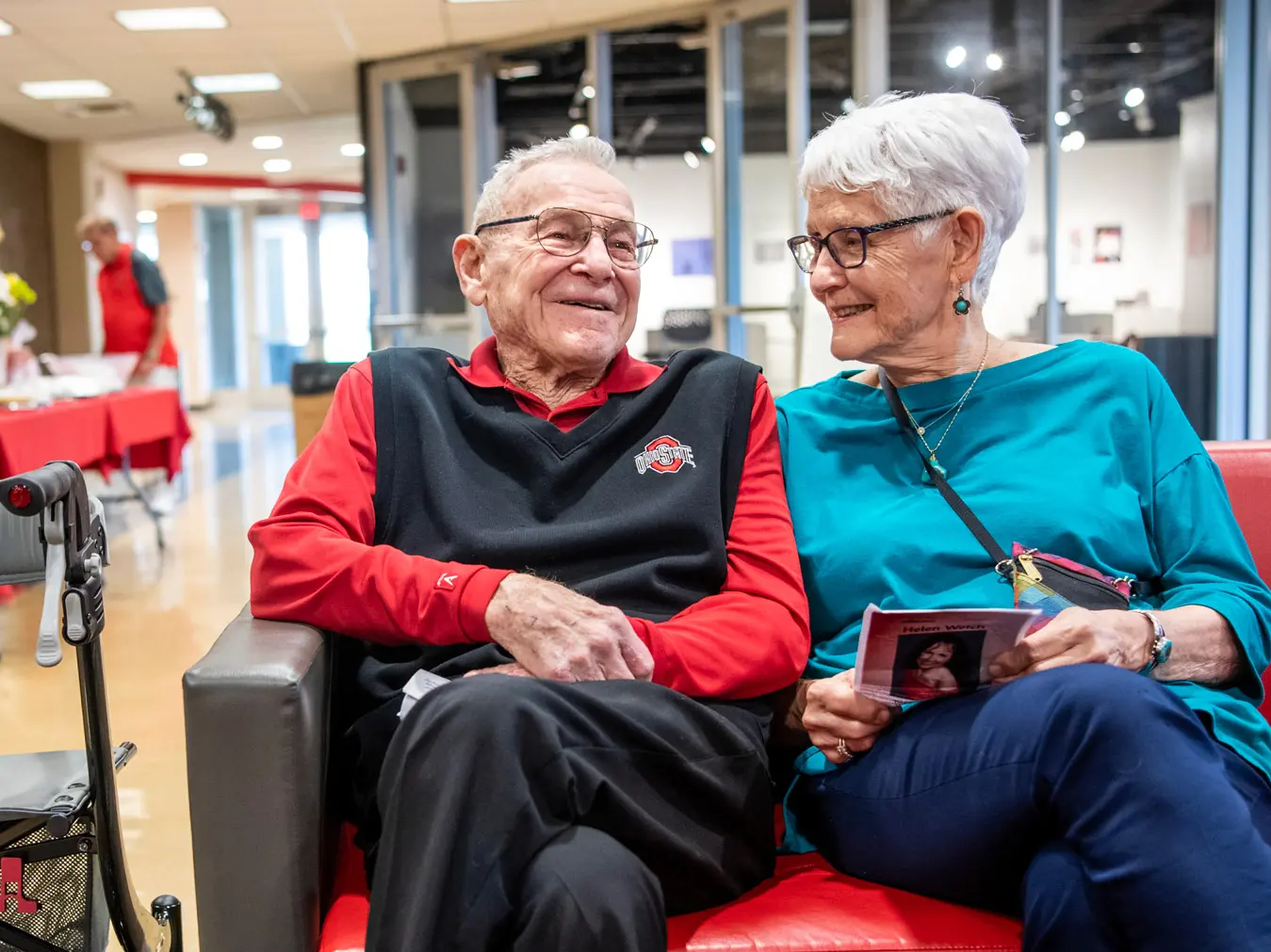 A white-haired couple sit together on a couch, smiling as they chat and lean against each other. 