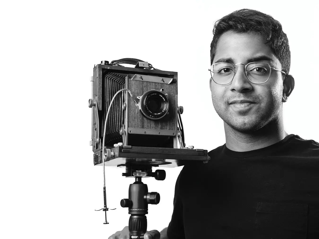 A young man of Indian descent stands beside a camera on a tripod and subtly smiles in a black and white self-portrait. He wears wire-rimmed glasses and his dark hair is short on the sides and a little curly on top.