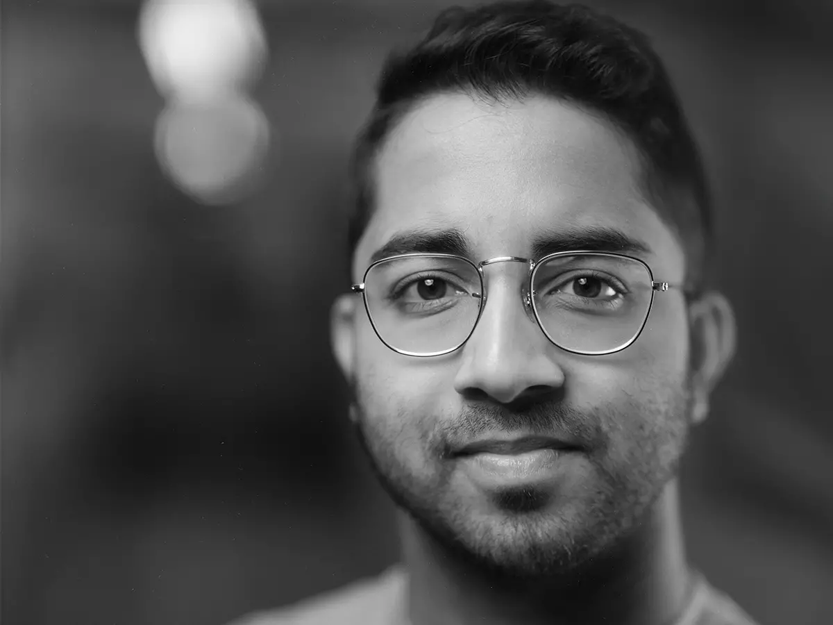 A young man of Indian descent subtly smiles in a black and white self-portrait. He wears wire-rimmed glasses and his dark hair is short on the sides and a little curly on top.