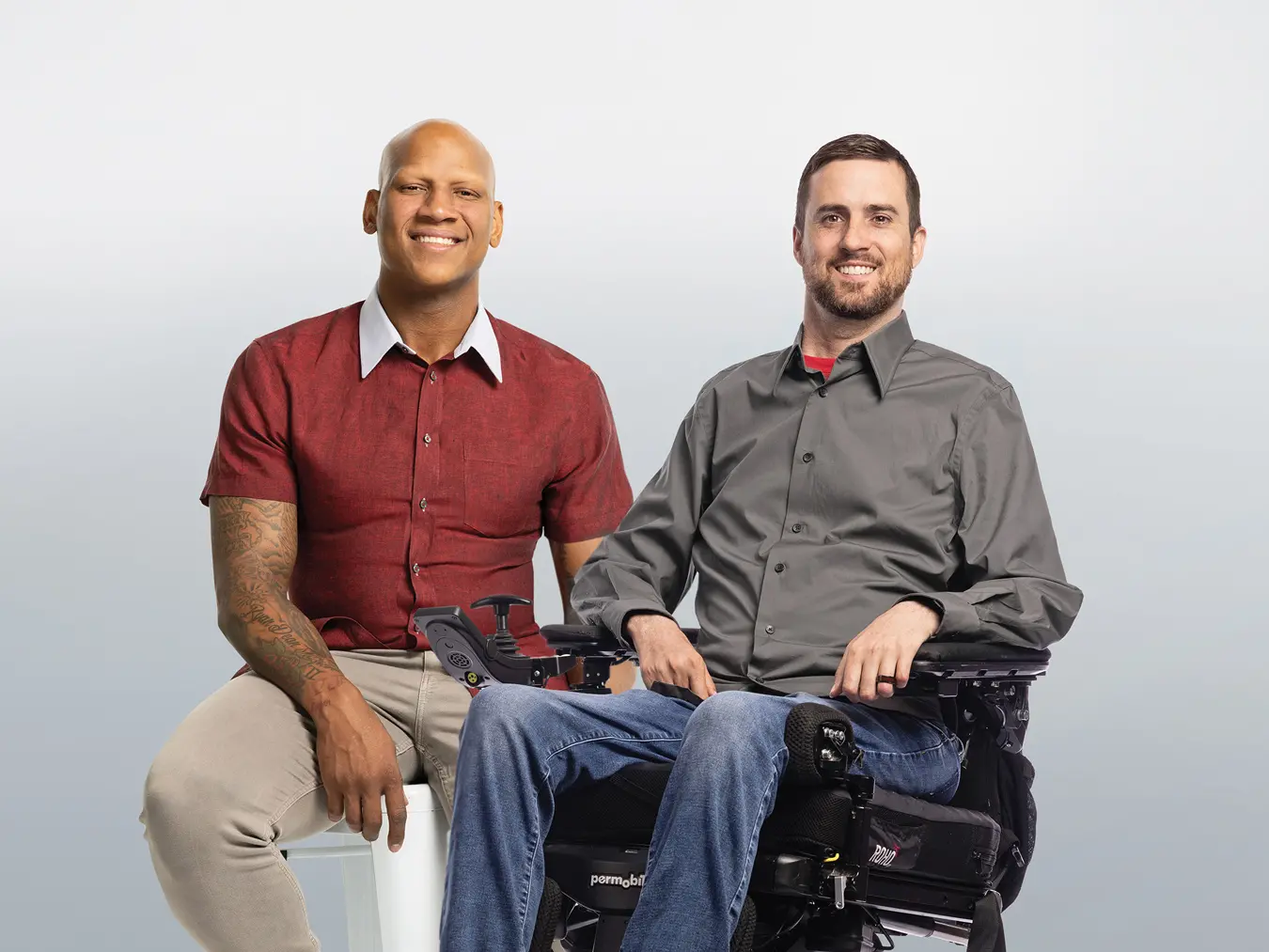 Two men sit next to each other smiling. On the left is a Black man on a stool wearing a short-sleeved button-down short that shows tattoos on his arms, a watch and shiny wedding ring. He casually rests his hands on his legs. On the right, a white bearded man in a motorized wheelchair wears a button-down shirt and a dark wedding ring.