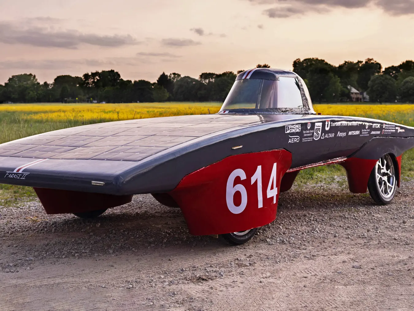The Ohio State team’s solar car is scarlet and gray. There’s a one-person cockpit and the main body is softly arch-shaped, coated in solar panels and surprisingly shallow. A large 614 is emblazoned on the wheel cover closest to the camera. The car sits in front of a field during a sunset. 