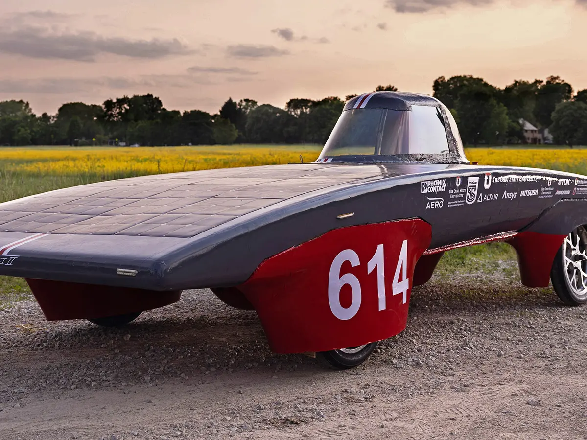 The Ohio State team’s solar car is scarlet and gray. There’s a one-person cockpit and the main body is softly arch-shaped, coated in solar panels and surprisingly shallow. A large 614 is emblazoned on the wheel cover closest to the camera. The car sits in front of a field during a sunset. 