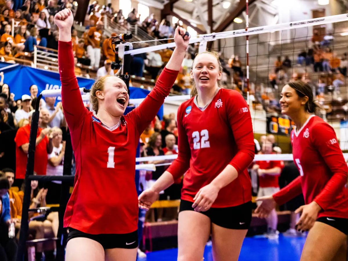 Three volleyball players wearing scarlet jerseys celebrate during a game in a gym as fans in the background cheer. A player with a No. 1 on her jersey is the most animated, with a huge grin and her hands fisted in the air.