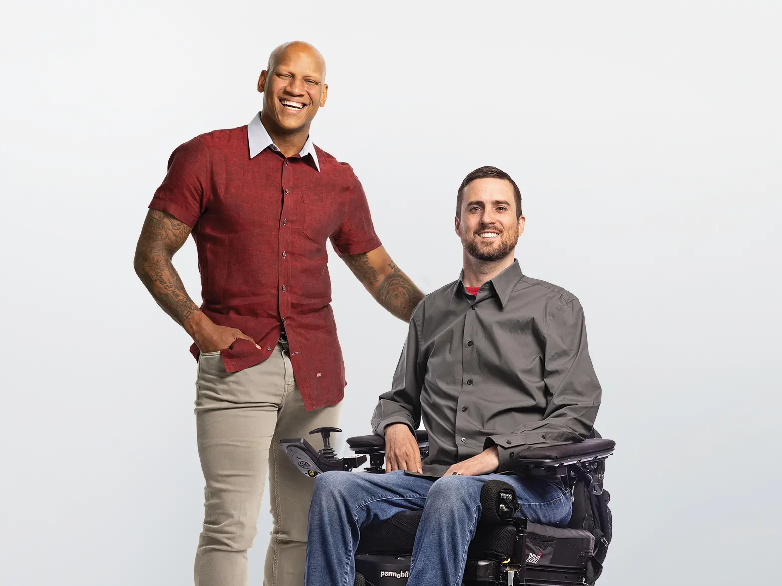 Two men are next to each other smiling. On the left is a Black man standing who is wearing a short-sleeved button-down short that shows tattoos on his arms. He has a hand on the back of the wheelchair of, on the right, a white bearded man. He wears a button-down shirt and jeans.