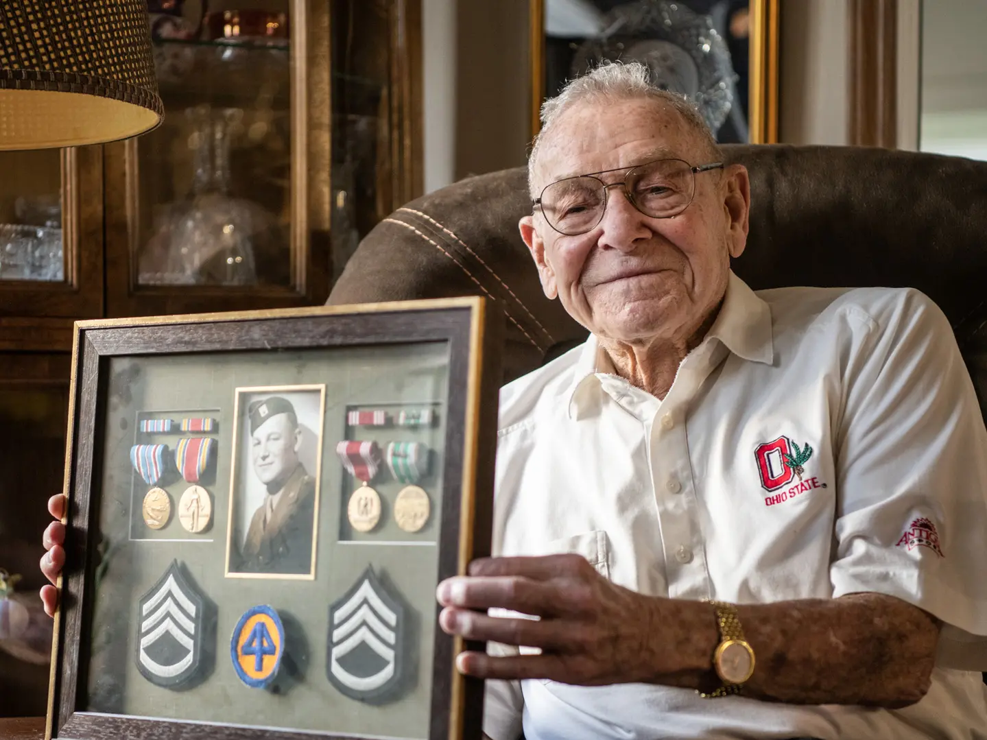 Merle Lashey Jr. shows a framed collection of military patches with his Army photo, taken when he joined to fight in World War II. He wears an Ohio State-emblem shirt and sits in a leather chair with a small, proud smile.