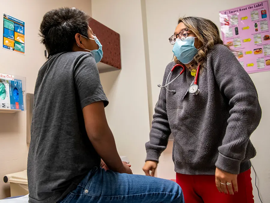 A young doctor in a surgical mask prompts a child patient to raise his shoulders during a medical exam