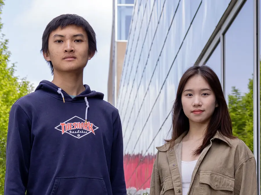 Two college students of Chinese descent pose for a photo next to Ohio State’s recreation building. A young man on the left wears a sweatshirt and sweatpants and a serious look. The student on the right is a young woman with long hair and a slight smile.