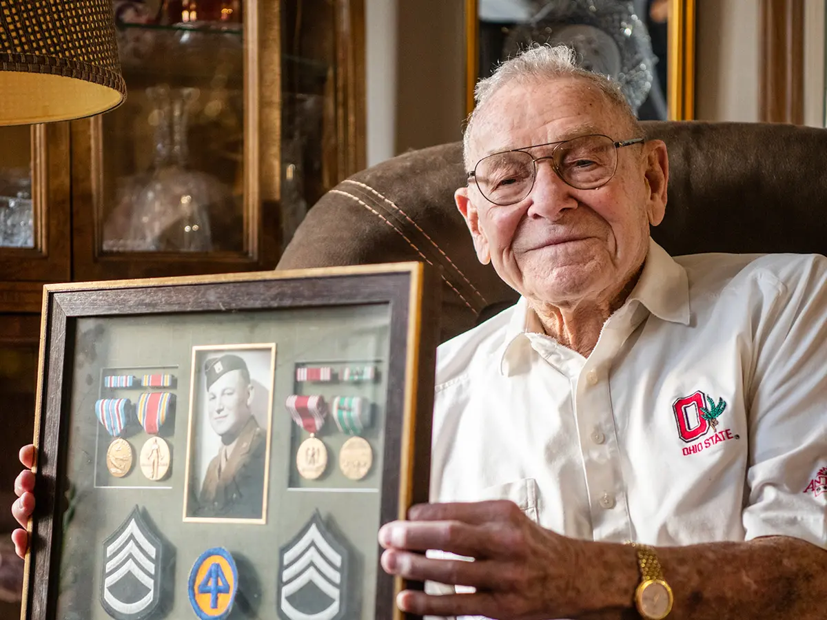 Merle Lashey Jr. shows a framed collection of military patches with his Army photo, taken when he joined to fight in World War II. He wears an Ohio State-emblem shirt and sits in a leather chair with a small, proud smile.