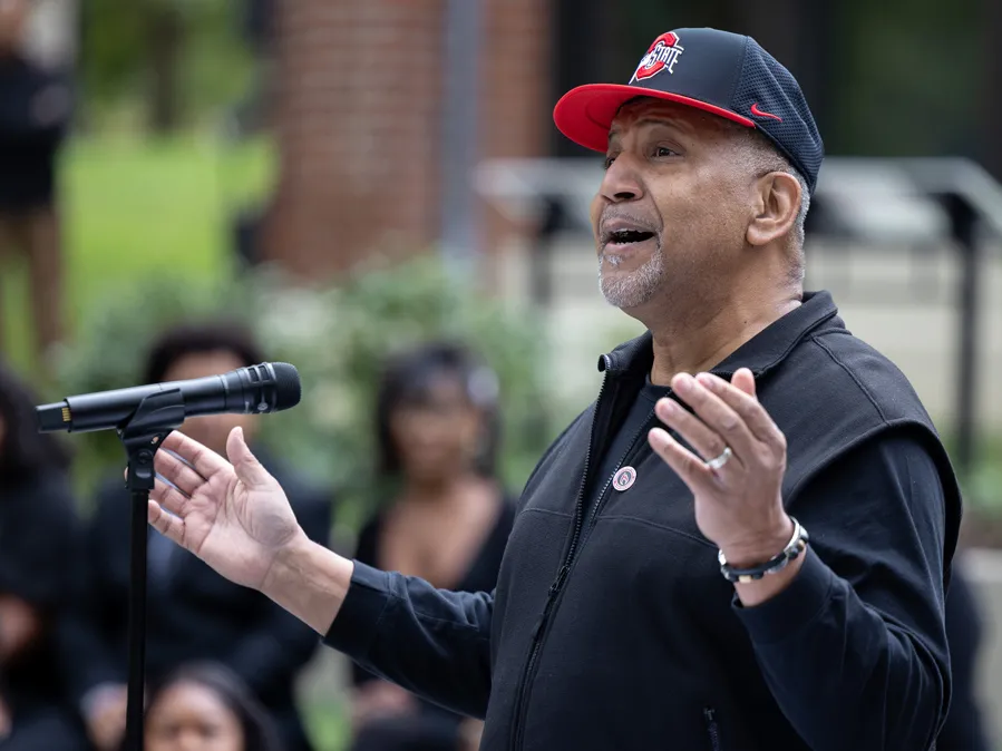 A Black man wearing an Ohio State ball cap gestures with hands spread wide as he speaks to an audience--only vaguely suggested in the background--at the dedication ceremony. He looks optimistic and proud. 
