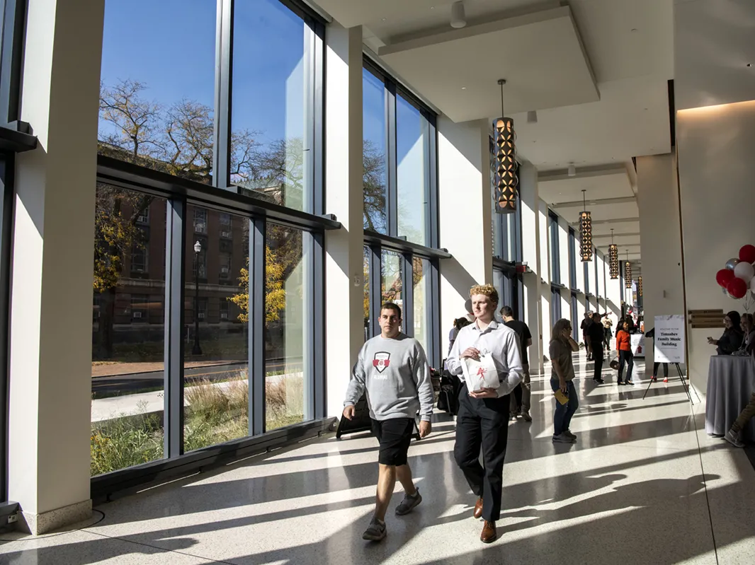 Students walk down a hallway where the late-afternoon sun streaming in through the wall of windows casts shadows on the floor.