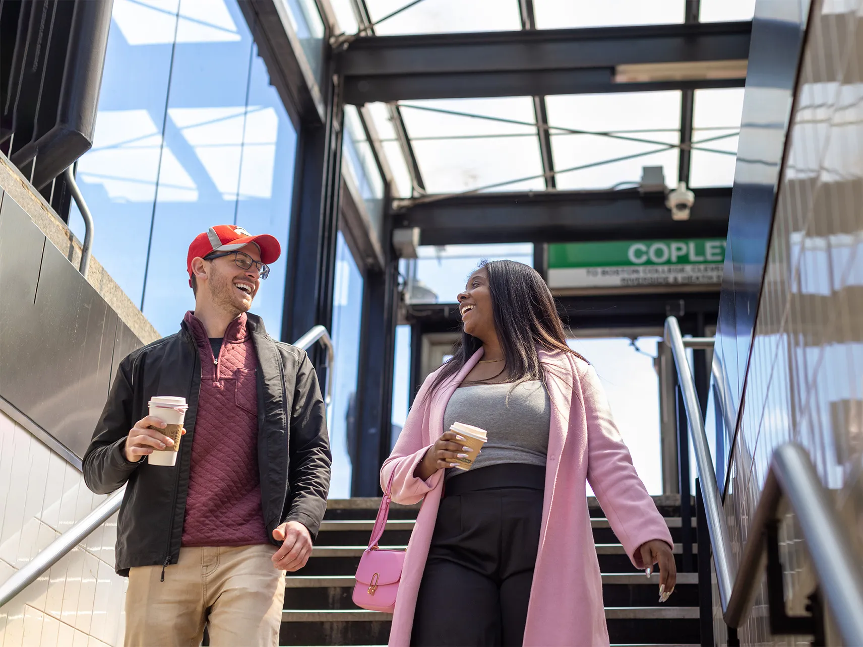 Josh Javor, a white man with glasses and a baseball cap, and Dominque McClean, a Black woman with long hair in a dress coat, smile at each other as they walk down stairs into a subway entrance.