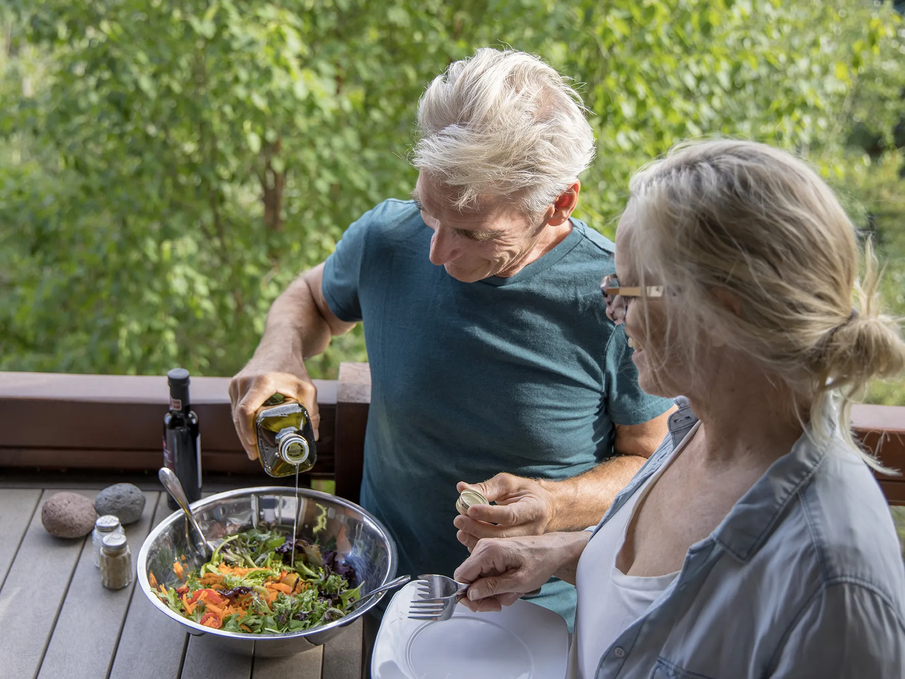Outside on a porch or deck, two older white people stand around a metal mixing bowl with a salad of greens, carrots and cherry tomatoes. The man is drizzling olive oil onto the salad and the woman holds plates and forks. Because the photo was taken from slightly above them and they’re both looking down at the salad, you can’t see their eyes.