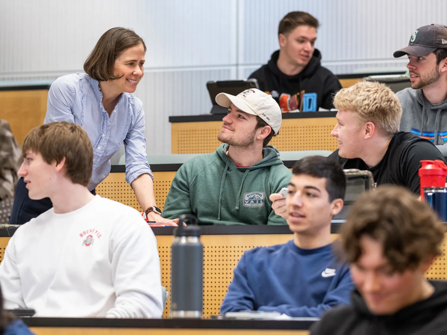 In a classroom with mostly male students, teacher Carmen Swain leans on a desk to talk to two students. She and the young men seem relaxed and are smiling.