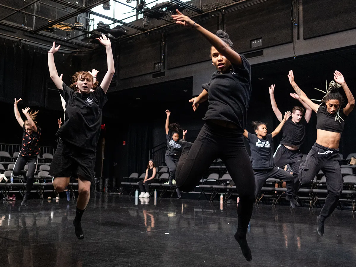 Seven dancers wearing black clothes—the color being the clothes’ only similarity—leap into the air with arms extended above their heads as they enact a scene from the island in “The Tempest.” 