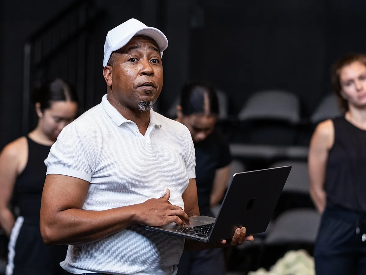 The choreographer of the class and chair of Ohio State’s Department of Dance, Charles Anderson, talks to the audience as his dancers stand in a line behind him. They’re dressed in black; his white shirt, gym shoes and ballcap stand out in the darkened theatre. He gestures and holds a laptop. 