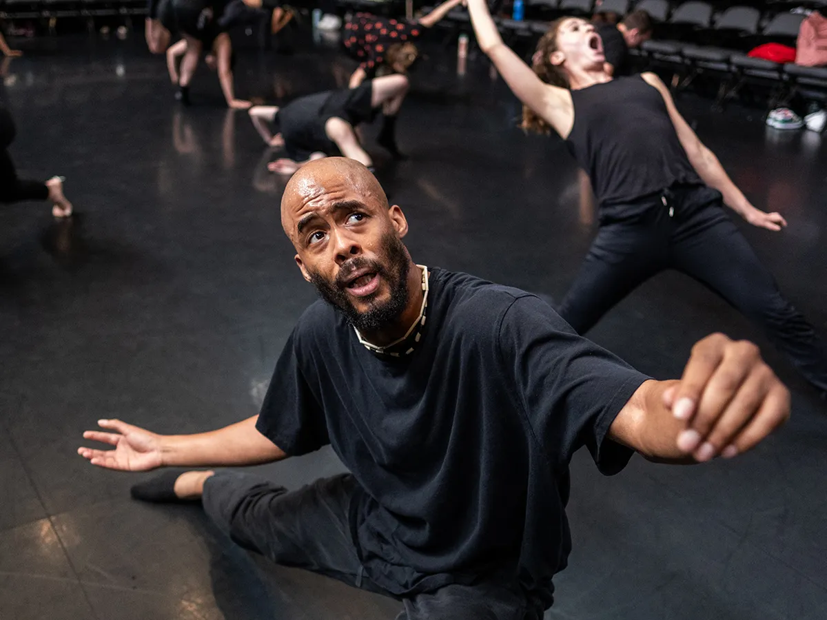 A man crouching, but with arms gently extended and his back leg stretched out, and looking to the ceiling appears perplexed or in awe. Sweat sheens on his forehead. Behind him, a dancer leans far back with mouth side open as she, too, looks above her. 