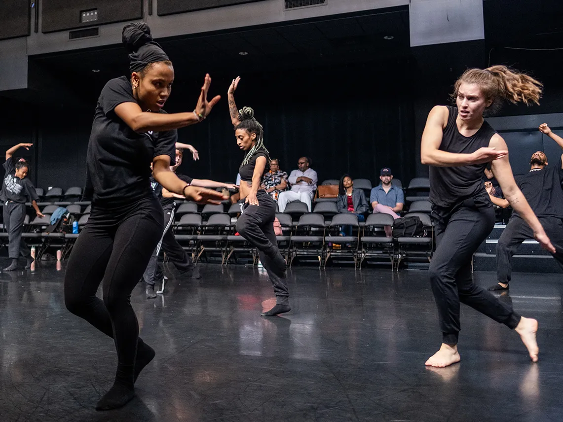 A young woman curls toward her left, looking that way and weight balanced on that side. Dancers alongside and behind her have limbs extended in various ways—they all seem very focused.