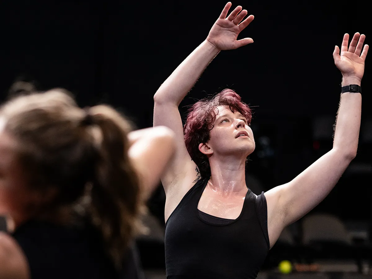 A dancer raises their arms, stretching toward the sky in a calmer moment of the dance. Their expression seems interested, curious and perhaps insightful. 