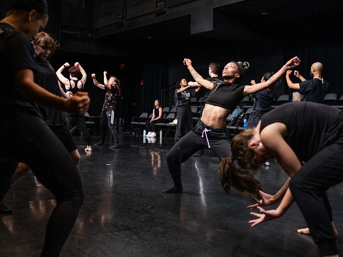 Four dancers perform in a loose circle on a dark stage. On the left, the two mostly stand, looking into the center of the circle. To the right, a dancer is hunched over looking at the floor, and the main focus in on the back right dancer. She leans back, arms stretched out, and looks to the sky.