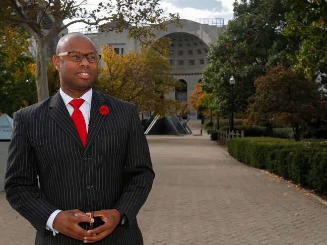 Dressed in a pinstripe suit and wearing glasses, a Black man stands in front of Ohio Stadium, looking into the distance and smiling