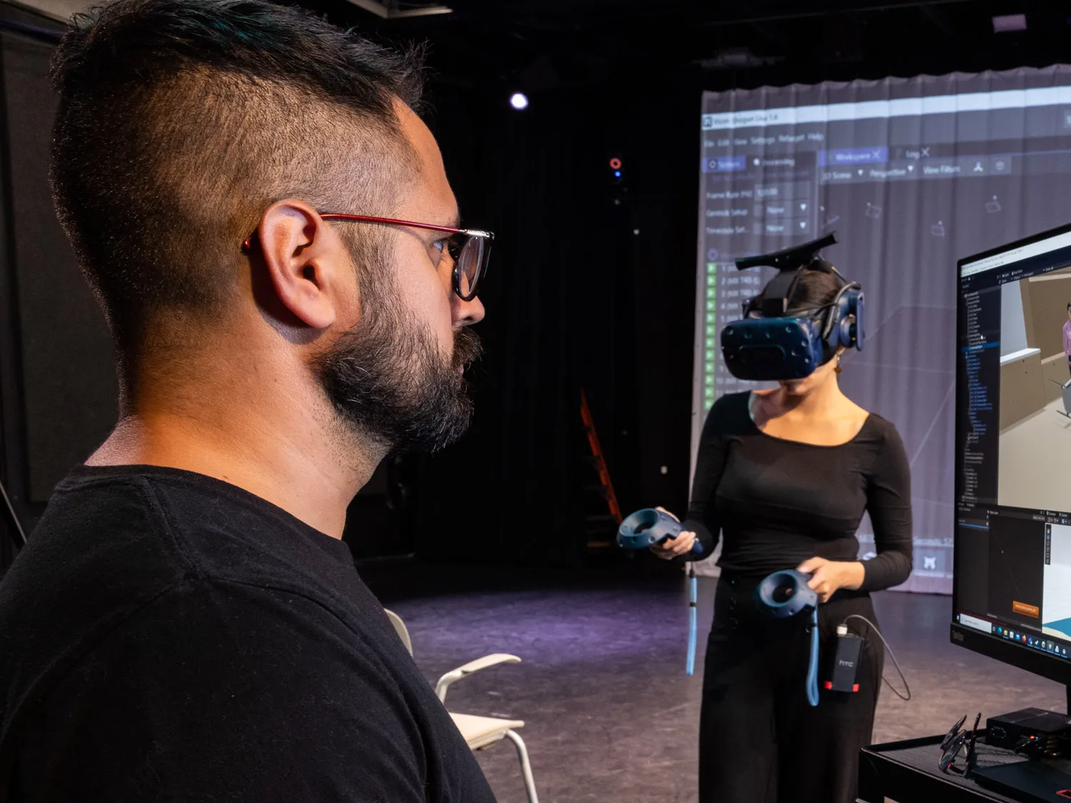 A man works on a computer whose screen shows a classroom setting while a woman on a stage beyond him wears a virtual reality headset and sensors used for motion capture. Ultimately, her movement will be shown on the screen he works on. 