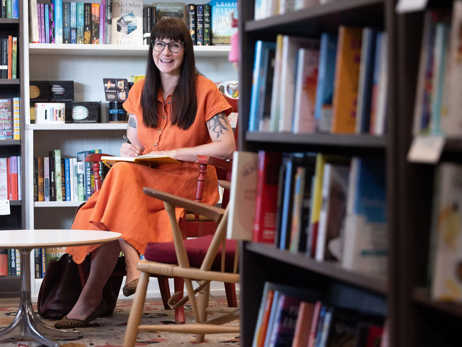 Maggie Smith — a white woman with long brown hair, glasses and tattoos of lemons and leaves on her upper arms, which you can see because she’s wearing a linen, short-sleeved dress —smiles as if she’s laughing or talking as she sits in a painted wooden chair. On her lap is a book she’s writing in and behind her are more books on shelves.