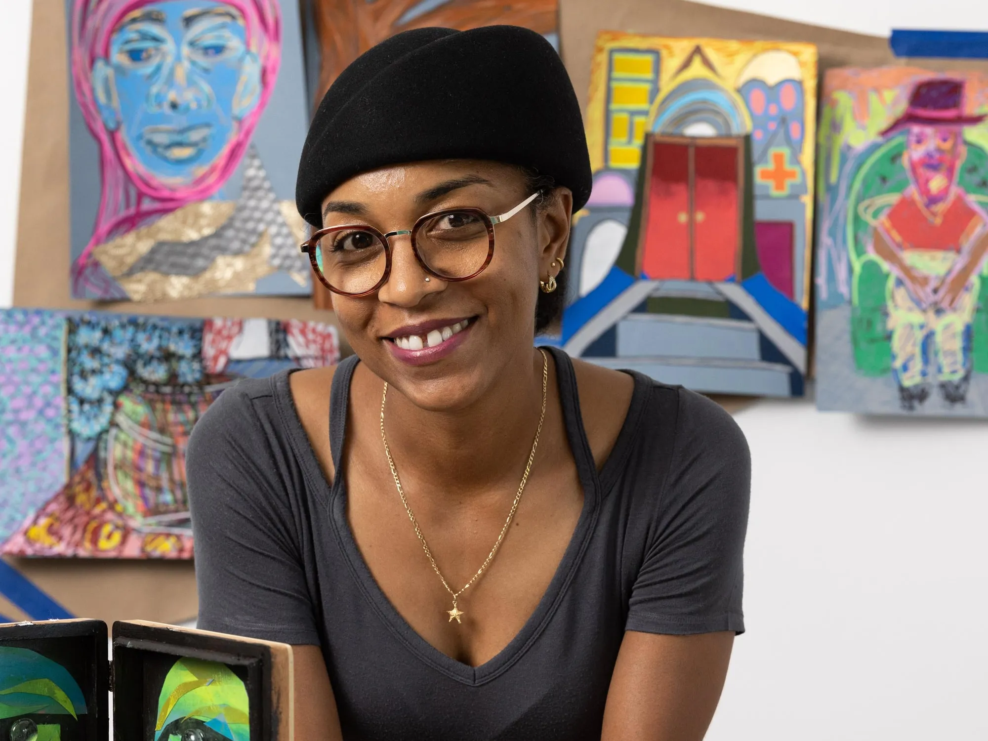 Tiffany Lawson, a Black woman wearing a hat, round glasses and a friendly smile, leans on her work table. In front of her are all kinds of supplies and works in progress and on the wall behind her are paintings she created on paper bags. They include people, faces and scenes in unexpected colors. 